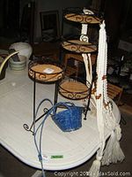 Photo showing black metal multi-tiered plant stand with wicker trays, three white macramé plant holders, small blue basket, and other pots on white table.