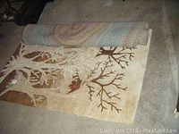 Unrolled rectangular area rug with nature/tree design in beige and brown colors on floor.