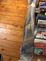 Rolled area rug in plastic wrapping placed on wooden floor near bookshelves