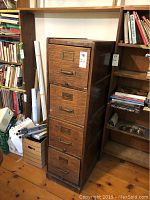 Front view of the tall vintage wooden 5-drawer filing cabinet positioned between shelves filled with books and boxes. Each drawer has brass handles and label frames.