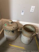 Full view of two pottery items on the floor against a beige wall: one large lidded jar and one double handled jug.