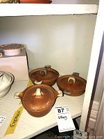 Three brown pottery bowls with lids displayed on a shelf, showing the shape, size and handles.