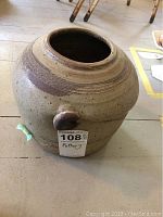 Large rounded pottery piece with a small knob on the side and textured horizontal lines near the rim in earthy brown and beige tones.