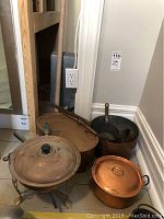 Photo showing assortment of copper pots including a chafing dish with stand and lid, a large pot, and smaller pots in corner on floor.