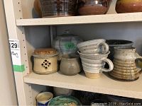 Shelf with various pottery items including jug, lidded jar with cork lid, stacked cups, small pot, and bowl.