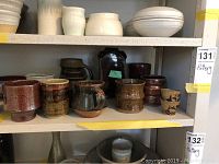 Photo of multiple pottery pieces arranged on a shelf including various cups, a jug, and a vase with earthy brown and white glazes