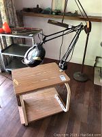View of wooden table with smooth light wood finish, lower shelf, and cream-colored metal support frame. Attached is a large, black magnifier light with an adjustable arm and circular lens. Table and magnifier placed on hardwood surface with natural light from windows.