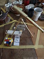 Photo shows vintage brass telescope on a tripod stand along with Rubik's Cube and light bulbs in front of various ceramics on a wooden table.