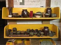 Two yellow shelves filled with assorted pottery items including bowls, vases, jugs, mugs, and small dishes arranged across the shelves