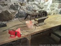 Photo showing a red bench vise attached to wooden worktable, terracotta pot next to it, and two anvils in background along stone wall.