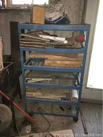 Photo showing entire shelving unit with wood pieces on second and third shelves, machinery, and hardware on top shelf, and miscellaneous items on bottom shelves.