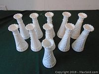 Front angled photo of 11 white milkglass bud vases showing their shape and decorative textures on a dark surface.