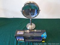 Photo showing a round make-up mirror with a metal stand and a boxed flat iron placed on a green cloth surface.