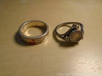 Two sterling silver rings displayed side by side on a wooden surface. One is a thick band with inlaid colored stones, the other features a round moonstone with decorative silver setting.
