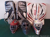 Front view of two pairs of goalie leg pads and three goalie masks arranged on a green surface.