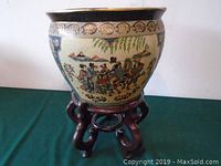 Full view of Asian ceramic planter on carved wooden stand, showing detailed painted artwork and planter shape.