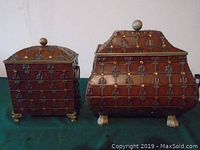 Front view of two metal decorative boxes with embossed fleur-de-lis pattern and brass or gold-tone accents, with curved feet and knobbed lids.