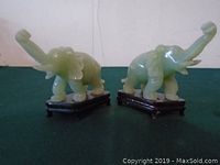 Front view side-by-side of two green jade elephant figurines raised trunks on dark wooden stands