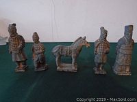 Five small Chinese warrior figurines and one horse figurine displayed on a green surface against a white wall background.