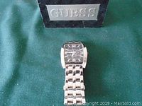Front view of Guess men's stainless steel wristwatch with rectangular black dial and silver markers, displayed on green fabric with Guess presentation box behind.