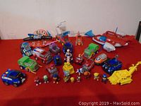 Full assortment of Paw Patrol toys arranged on red carpet, showing a variety of vehicles, figurines, and electronic toys.