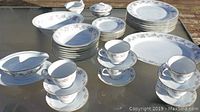 Wide view of entire 45 piece RC Chantilly dinnerware set arranged on glass table showing cups and saucers stacked, plates in piles, serving platters, covered sugar bowl, and gravy boat.
