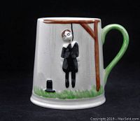 Front angled view of the ceramic mug displaying the raised hangman figure hanging from a brown gallows structure with a green handle and a black top hat on the grass below.