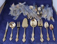 Gold plated flatware pieces arranged on blue background, showing multiple utensils including various spoons, forks, ladles, and serving pieces. Pieces are used but well maintained.