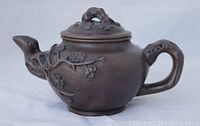 Side view of antique brown clay teapot showing curved handle with floral relief detail and bird-head shaped spout.