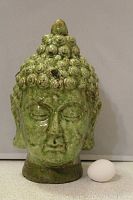 Front view of the vintage green pottery Buddha bust with a green glaze and textured curly hair detail next to an egg for size reference.
