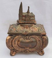 Front and side view of ornate copper wash metal trinket box with detailed decoration of a church and nature scene on top.