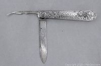 Silver tone antique/vintage pocket knife with one straight blade and one curved hooked blade extended, showing textured handle with embossed circular floral motifs on silver tone metal.