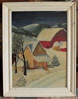 Framed winter landscape painting showing houses, barns, snow and hills in folk art style with white frame.