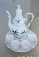 Miniature white porcelain teapot with curved handle, spout, bamboo leaf hand-painted motif and gold accents, displayed on matching tray with four teacups.