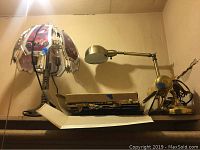 Three table lamps placed close together on a shelf, including a metallic adjustable lamp, a decorative purple-pink lamp, and a motorcycle-themed touch lamp with glass top. Stack of stick-on mirror squares partially visible.
