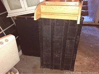 Two black steamer trunks side by side against a wall with wood paneling and miscellaneous items around, showing size difference and metal corner details.