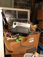Cardboard box filled with various vintage toys and electronics. Visible is a portable radio resting on the top of the box, along with toy parts and other items. The box is surrounded by clutter and appears dusty and worn.