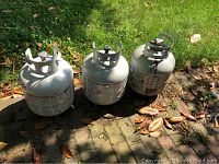Three propane tanks placed outdoors on a stone surface, viewed from above. Tanks show signs of outdoor exposure, valves and top collars intact.