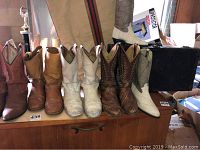 Multiple pairs of men's boots lined up on a wooden surface including brown, tan, and white boots with western styling and decorative stitching.