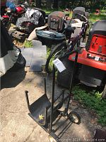 Black metal firewood dollie standing on concrete surface outdoors near various outdoor equipment.