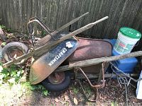 Two wheel barrows placed on grass near a wooden fence, both showing rust and wear, one branded Craftsman