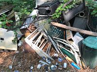 Pile of heavily rusted scrap metal with pieces like ladder sections, chrome bars, rusted sheets, green wire mesh basket, wood plank, and loose debris scattered on ground with some vegetation