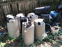 Photo showing large beige plastic trash cans with wheels upside down on grass, some with black lids and others partially covered by debris.