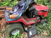 Side view of the red Murray riding mower showing worn seat with tarp cover and bricks on it, engine compartment, front and back tires, and detached plastic cover on ground.