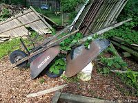 Two metal wheelbarrows shown side by side outdoors on dirt and grass with wooden debris nearby; one has AGRISTAR logo and damaged frame.
