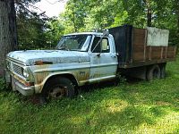 Side view of 1970 Ford F350 dump truck showing rusted body, wood dump bed, flat tire, and weathered paint.