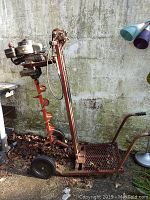 Side view of the gas powered post hole digger showing the motor, red auger, metal frame with mesh platform and wheels.