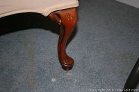 Close-up photo of one wooden curved leg of the tan armchair on carpeted floor.