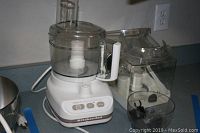 Full view of KitchenAid food processor with attached clear bowl, lid, and additional attachments visible on the side.