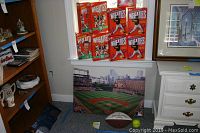 Room corner with four Wheaties boxes stacked on windowsill, framed Camden Yards panoramic photo below, signed football on floor.